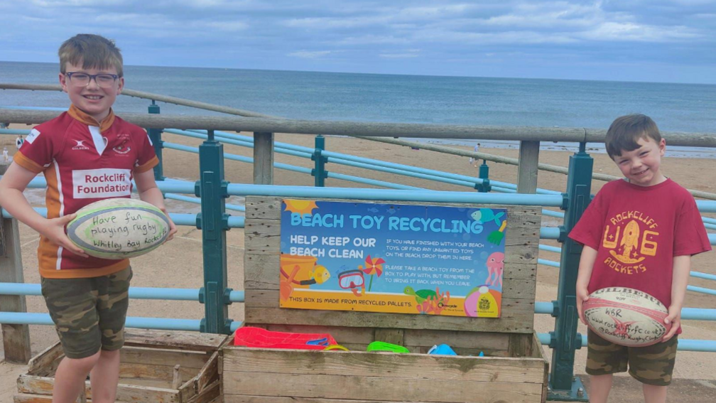 Two children both holding rugby balls at Longsands beach. They are standing between a wooden chest, where people can donate toys for other people to use.