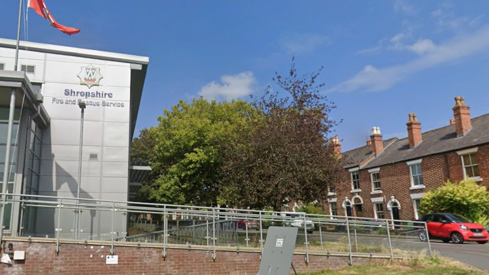 A silver fire and rescue service building with a sign that reads "Shropshire fire and rescue service". there is a red flag flying on a pole outside it. To the right of the building is a road with traffic queueing