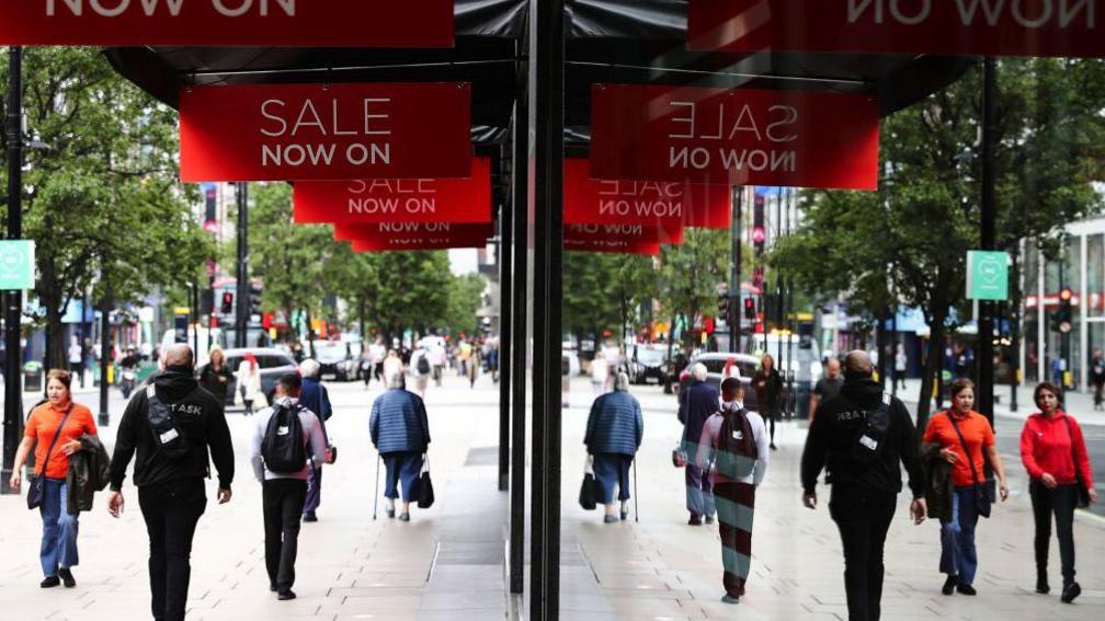 Pedestrians pass sales promotion signs outside a House of Fraser department store, operated by Frasers Group Ltd, on Oxford Street, London, in July 2020. The pedestrians and signs are reflected in the shop window. 