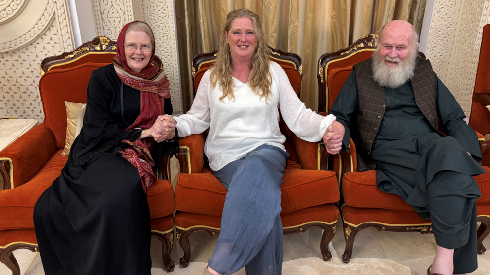 Freed British couple peter and barbie Reynolds sit with their daughter sarah. On the left, Barbie is wearing a dark dress. A blond haired Sarah is sat in the middle of the two, wearing blue jeans and a white top. On the right sits Peter, he's wearing a traditional Afghan dark top and trousers, with a brown textured waistcoat. All are smiling at the camera.