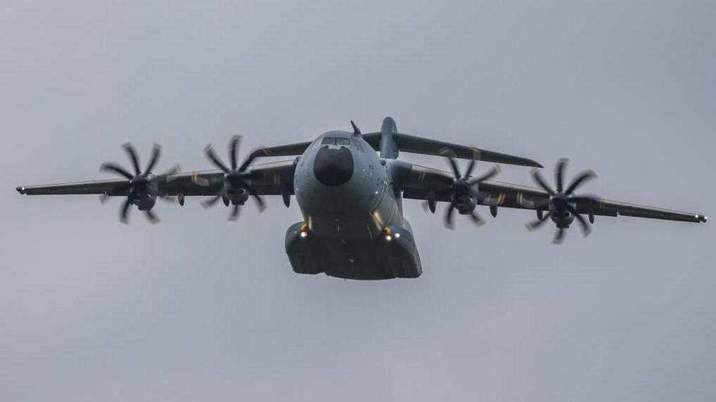 An Atlas A400M aircraft. The plane has four propellers and a broad grey body and raised tail and is in flight in the picture with the wheels raised up 