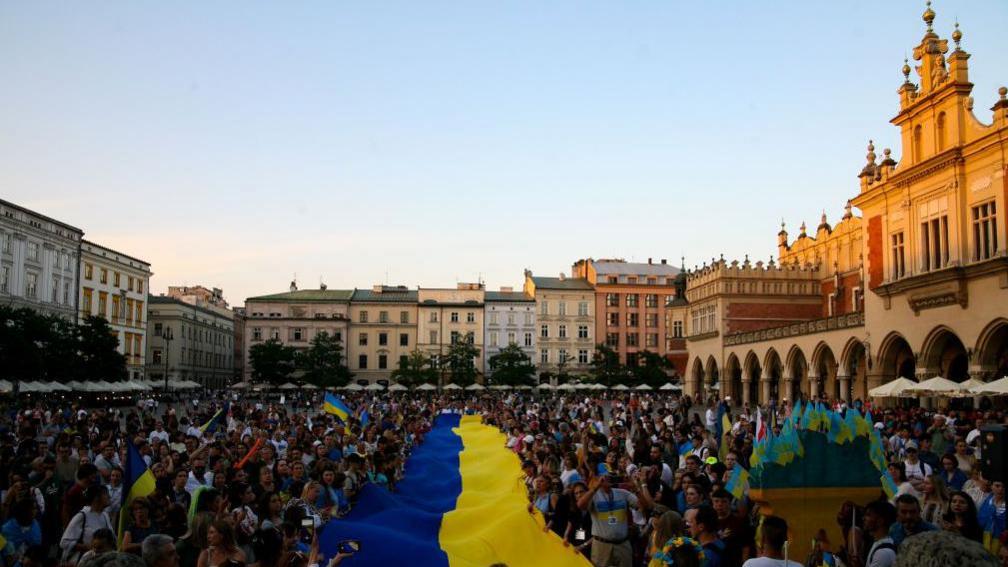 A large crowd of people in a square surrounded by buildings on three sides. A large blue and yellow Ukrainian flag is displayed running through the centre of the crowd. 24 August 2023