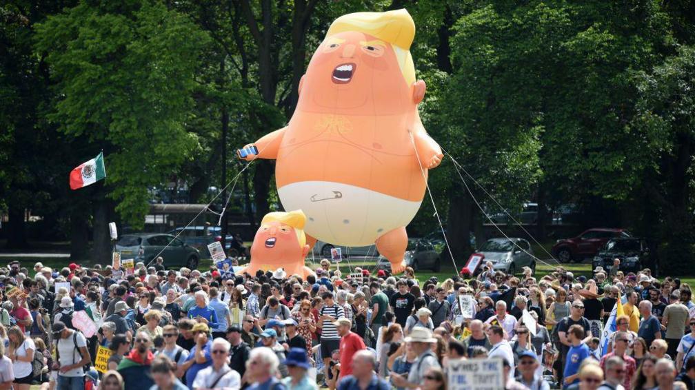 A photo of a protest in Edinburgh in 2018. There are hundreds of people milling around but the photo is dominated by a huge inflatable baby Donald Trump wearing a nappy