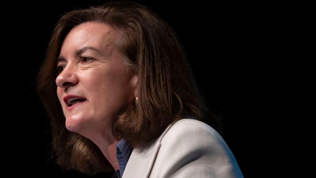 A head and shoulders photograph of Eluned Morgan giving a conference speech. She is looking towards the audience, away from the camera, and wearing a blue blouse and white jacket