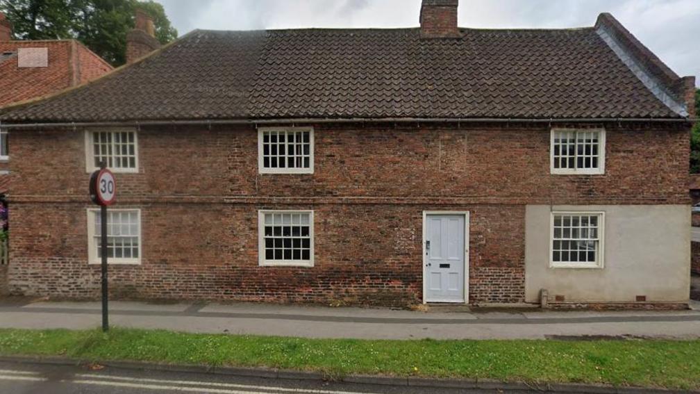 An 18th Century steep roofed building with small windows and a light blue door sits in front of a footpath and strip of grass