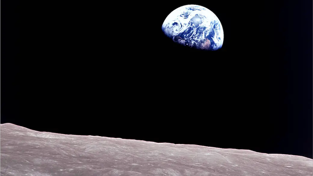 A three quarters view of the Earth appearing as a blue marble with white swirls, rising above a lunar landscape