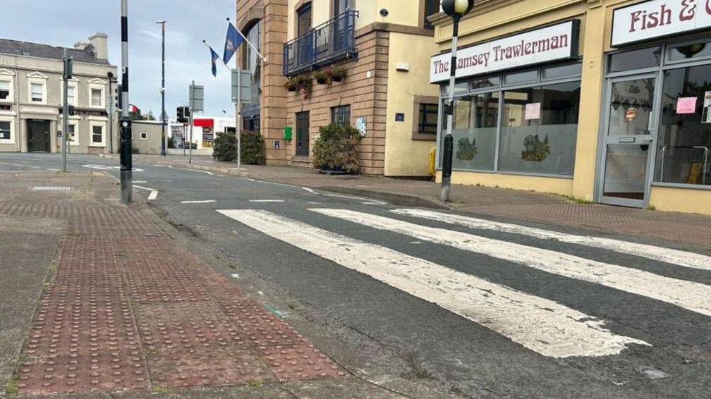 A section of road in Ramsey in front of the town hall, which sits next to a chip shop. You can see tired-looking pavements on both sides of the grey tarmac.

