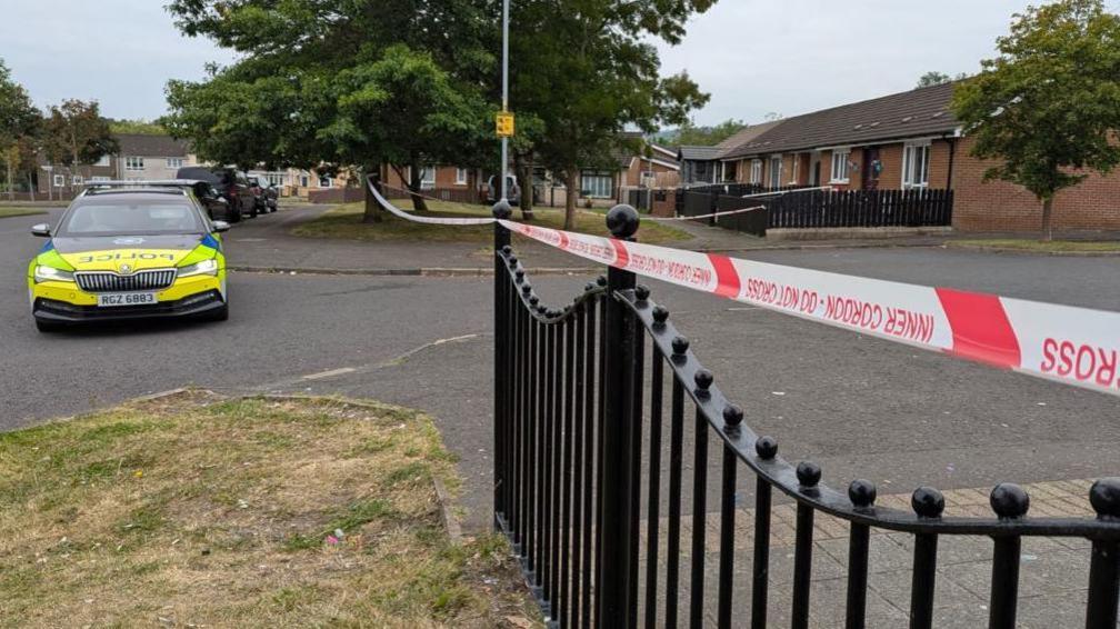 A police car which is yellow and blue in colour is parked to the left of a police cordon tape. Police is written on the side and front of the car in blue letters. To the right of the picture are houses and to the fore are black railings with the police tape across them.