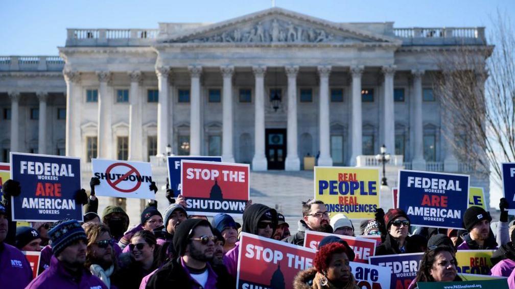 People attend a rally of air traffic control and other airline industry unions on Capitol Hill in Washington, DC, on 10 January 2019 during the previous US government shutdown. People hold placards reading "Federal workers are American workers", "Stop the shutdown" and "Real people, real consequences".