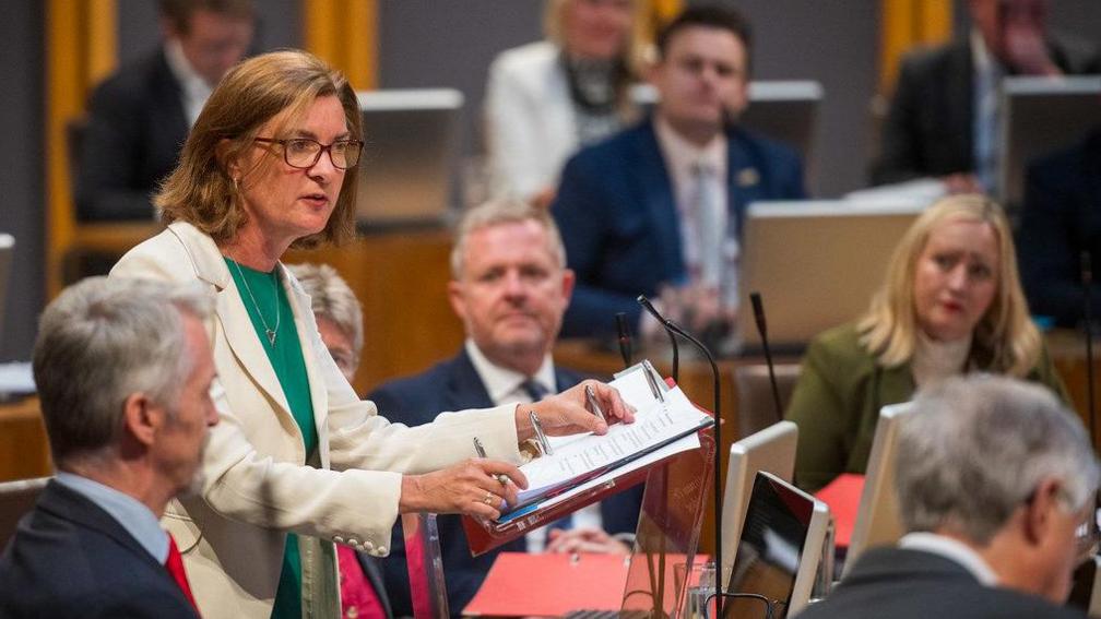 Eluned Morgan on her feet answering a question in the Senedd holding a binder and a pen and surrounded by ministers listening closely to her words  