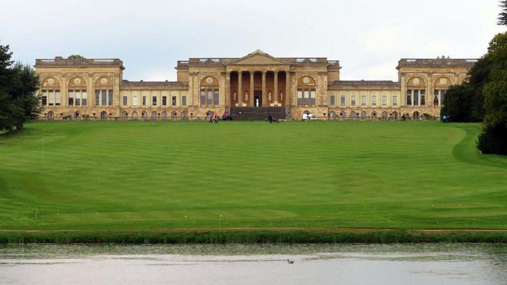 Stowe School with the grass field in front. It is an imposing neoclassical building with pillars at the front entrance.