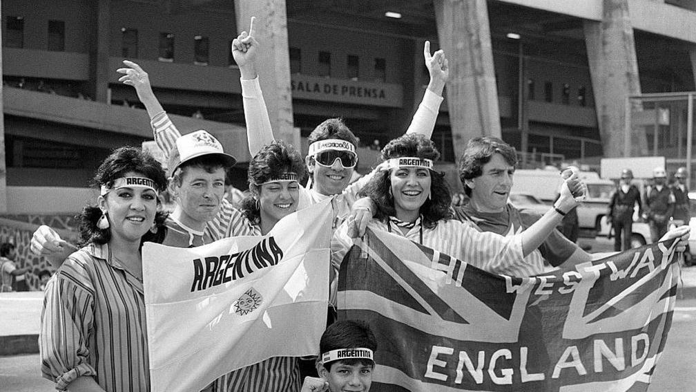 English and Argentine fans share a laugh and a joke outside the Azteca stadium. The photo is black-and-white and they are holding banners from their respective countries and smiling broadly. 