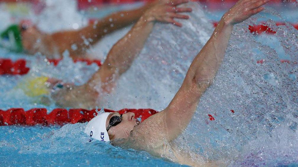 a swimmer doing the backstroke, his white cap and black goggles peak out from the water, his arm is extended and surrounding by water splashes.