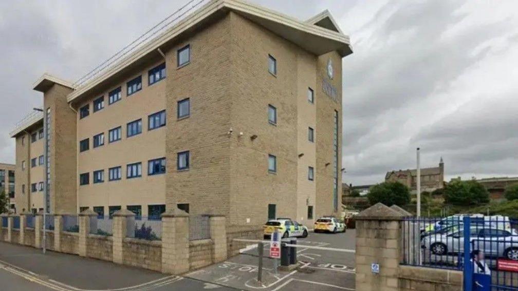 A beige four-storey building with a car park which is barriered off. Police vehicles are parked in the car park.