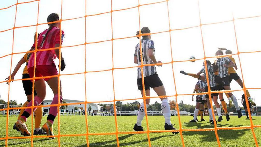 A shot taken from the back of an orange football net. Several players have their backs to the camera and the ball is in play. Some of the players are jumping in the air to head it.