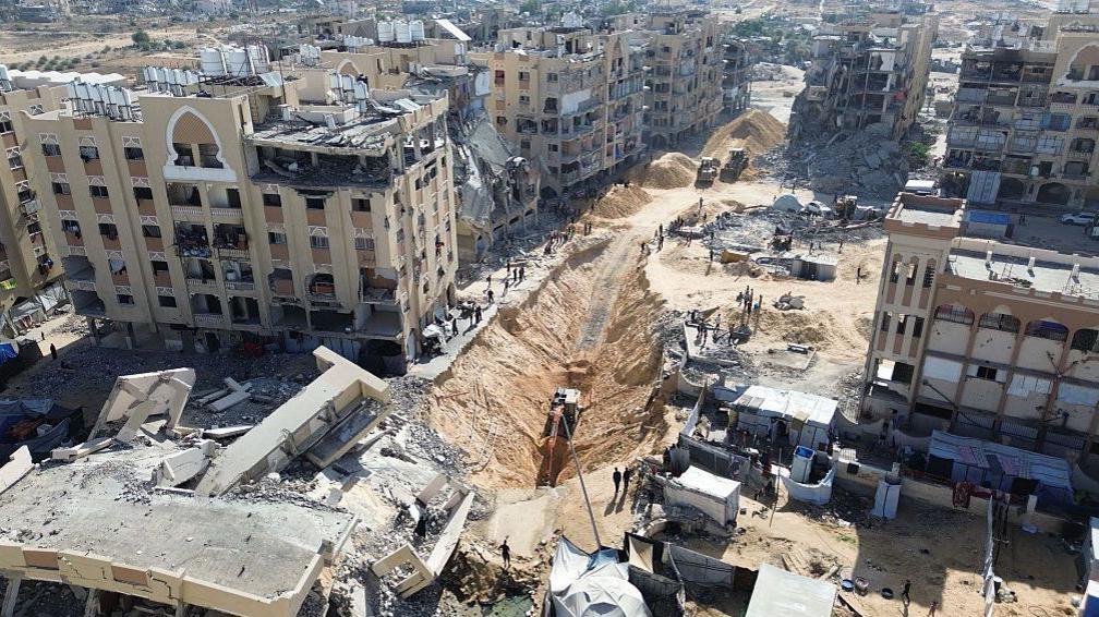 Excavators dig a trench amid damaged and destroyed buildings in the Hamad City neighbourhood of Khan Younis (21 October 2025)