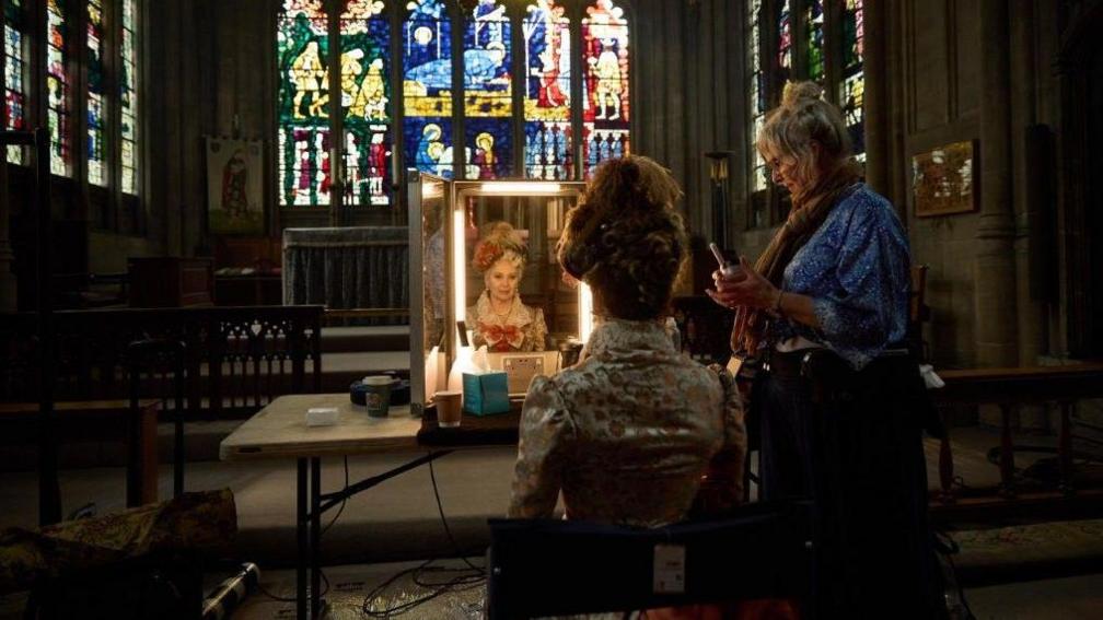 A woman in period costume sitting in front of a dressing table mirror. She has her hair done up and is wearing a small bonnet with feathers, as she looks at her reflection. To her right is a woman holding a walkie talkie. In the background can be seen a church altar beneath three large stained glass windows.
