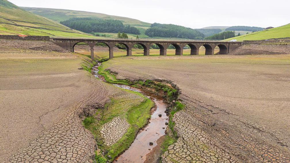 view of a dried up reservoir with cracked and brown ground along with a small stream running through it with a bridge in the distance