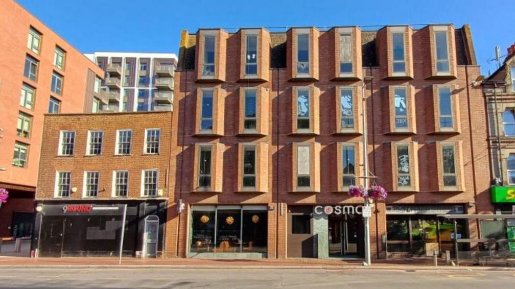 A photograph of the area as it is now. It's a red brick four-storey building, with a 'Cosmos' restaurant sign on the ground floor. Next to it is a three-storey, flat-roofed townhouse with a sign for '9 Round', a kickboxing gym. It's a sunny day and tower blocks can be seen next to and behind it.