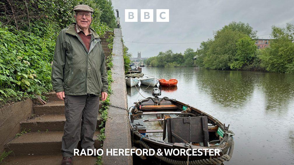 Man in a flat cap and green jacket standing on concrete steps leading to a boat on the river.