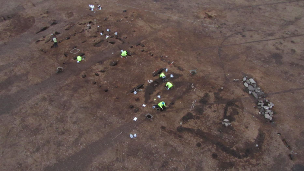 An aerial image of the dig site. The ground is bare, brown earth and archaeologists wearing fluorescent jackets work at uncovering finds. The long "footprint" of the hall is evident.