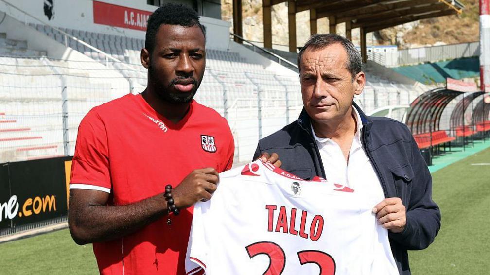 An Ivory Coast footballer in a red football shirt stands with an older man to the right while the two of them hold a white shirt labelled Tallo 22