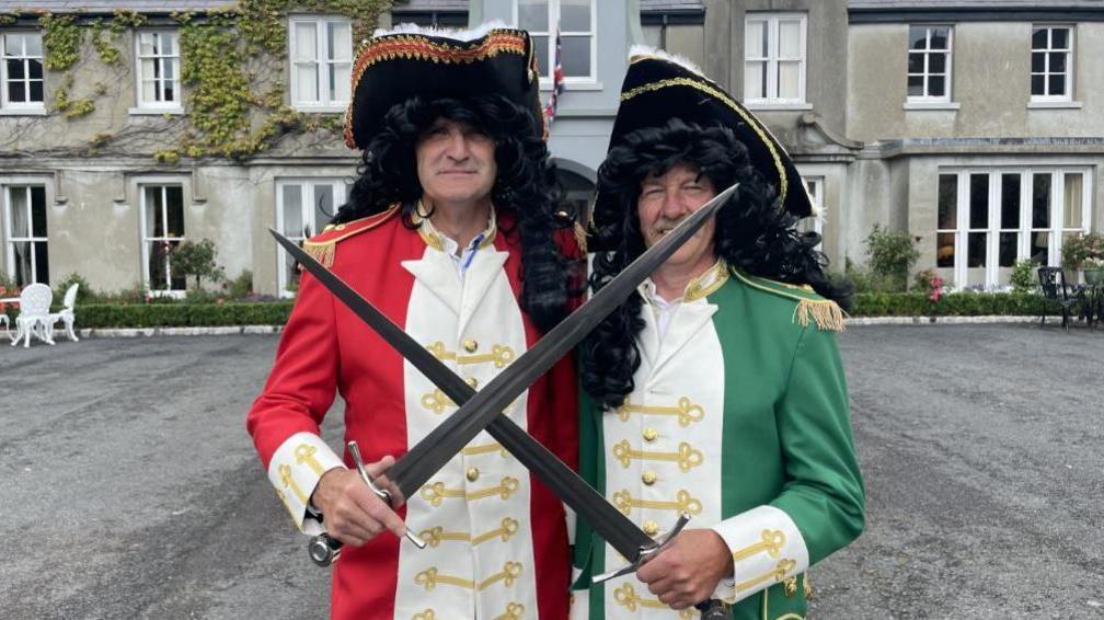 Two actors dressed as King William III and James II. William is dressed in red and white and James in green and white. Both are wearing long black wigs and are crossing swords. 