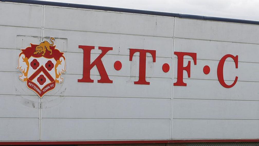 A Kettering Town club crest and letters spelling KTFC (Kettering Town Football Club) on the side of a stand at Nene Park. The faded outline of Rushden and Diamonds' badge and the letters RDFC are faintly visible beneath the new signage.