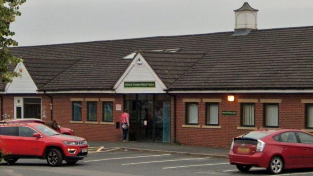 The front of a one-storey red brick building with two red cars outside and a green sign above the entranceway