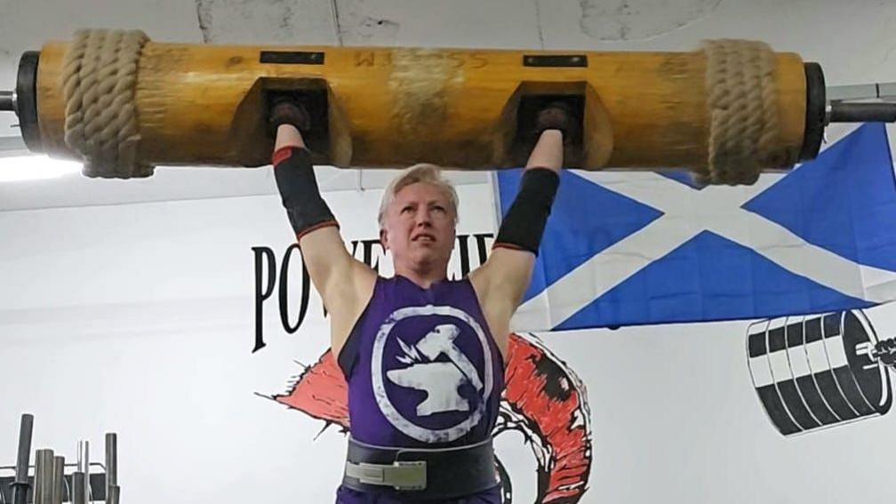 A woman in a gym pressing a heavy wooden and metal barbell over her head. She wears a purple vest, elbow supports, and a thick leather belt around her waist