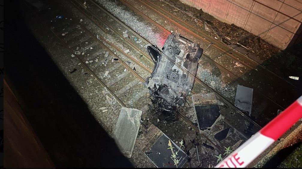 A burnt-out and smashed BMW lies on its roof on the railway line. Debris is scattered all around the wreckage. The photograph was taken at night, with only the car wreckage illuminated.