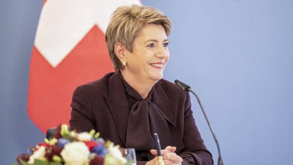 Switzerland's president Karin Keller-Sutter smiling sat at a desk with a blue background and part of a swiss flag behind her