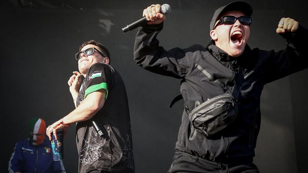 Kneecap performing on stage. One wearing a black jacket and baseball cap. One wearing a black and green t-shirt and one wear a green, white and gold balaclava.