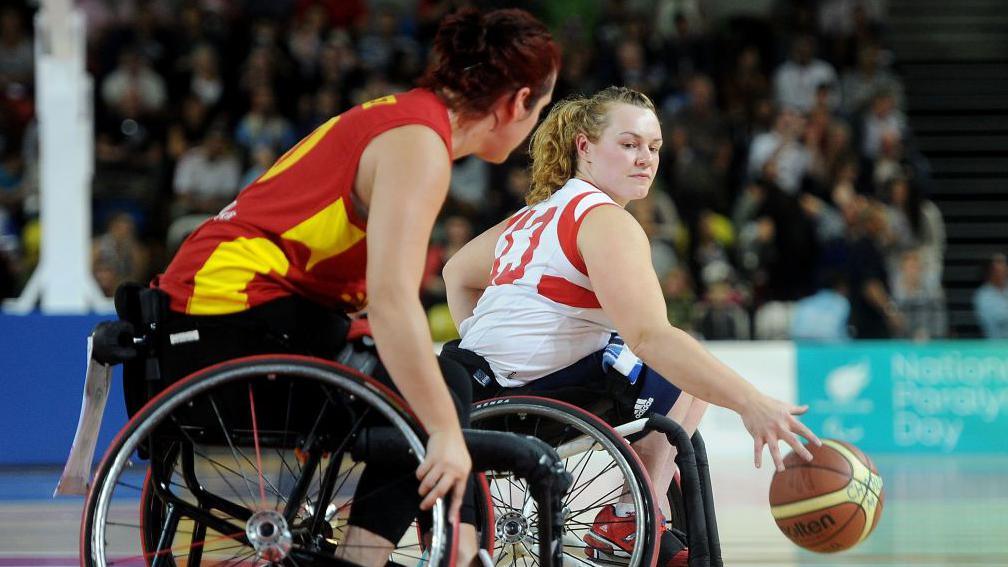 Louise Sugden plays Wheelchair Basketball for Great Britain. She currently has the ball, and is looking over her shoulder as a member of the opposing team approaches. 