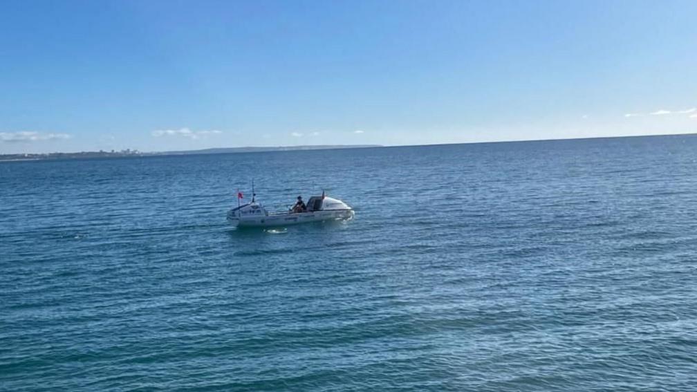 Zara Lachlan in a white wide-beamed 24ft (7m)-long boat. She is rowing on blue ocean water, which is calm, with paler blue sky above