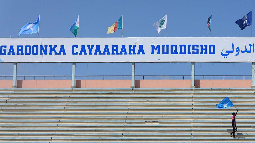 A lone figure waving a Somalian flag above his head walks through the empty stands of a stadium. Above him, the flags of various African nations fly above a white sign with "garoonka cayaaraha mogadishu" (Mogadishu Sports Stadium) printed on it in blue block capital letters.