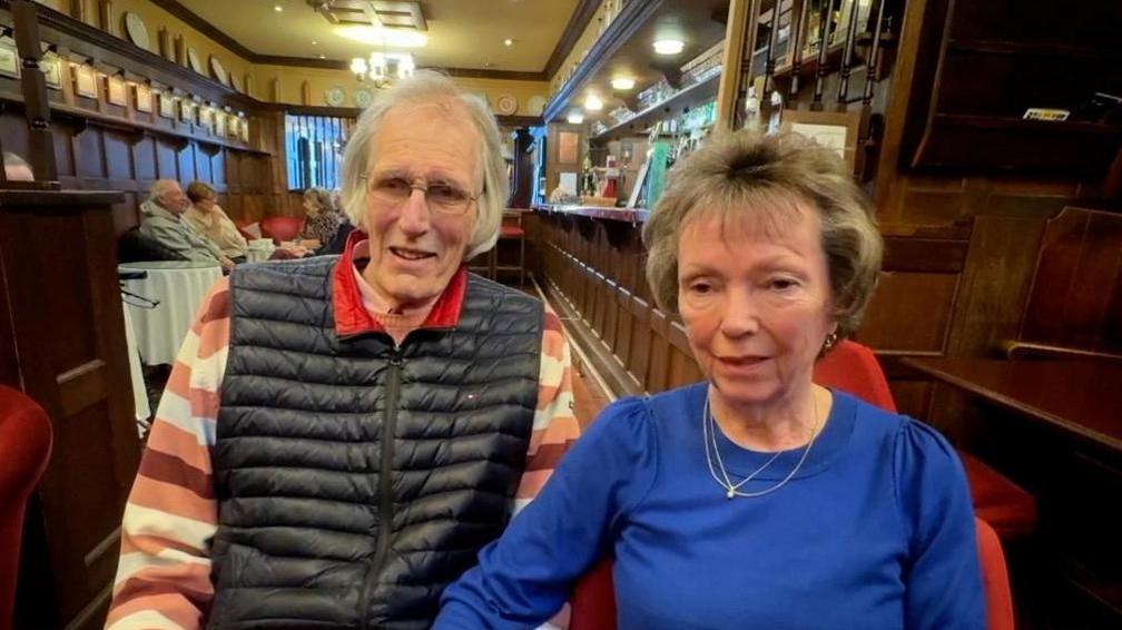 George and Rita Riggs sit in the oak panelled bar in their hotel. Customers are visible in the background. Mr Riggs is on the right of the image and has long grey hair and glasses. He is wearing a navy blue gilet with a striped jumper. Mrs Riggs has grey hair and is wearing a royal blue top with gold jewellery.