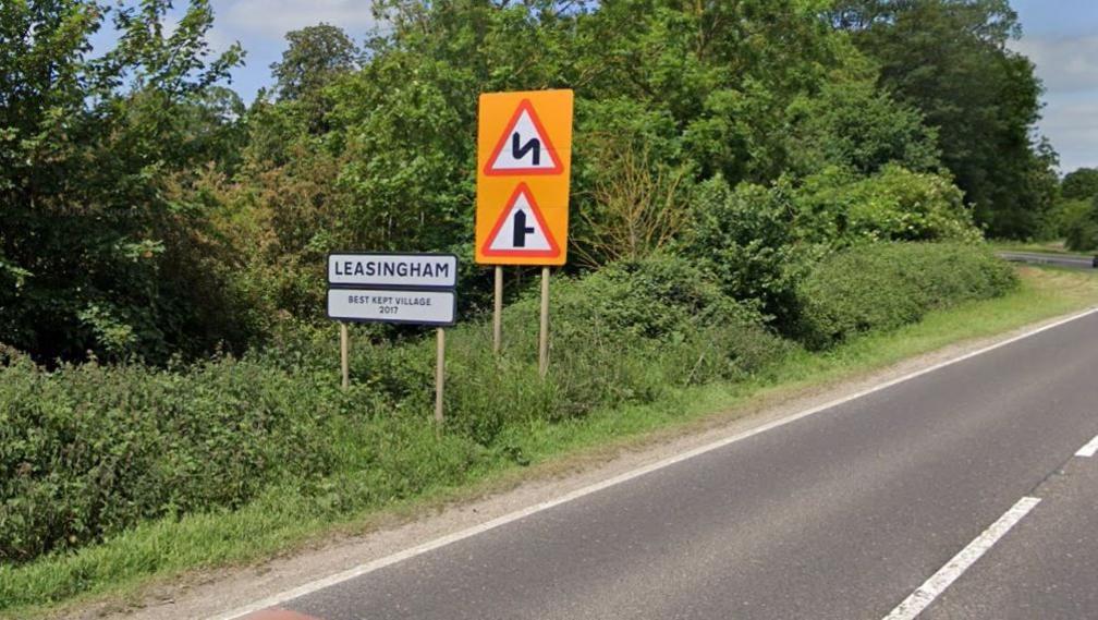 Black and white sign for Leasingham on the side of the main A15 road, with two red triangle signs behind it - one denoting a junction - the other bends in the road