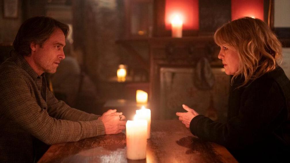Ashley Jensen's character DI Ruth Calder is sitting at a wooden table with Stuart Townsend's character Ed Tulloch. On the table are two large candles which are casting a warm glow over the pair. 
There are more candles in the background along with a fireplace or mantel.