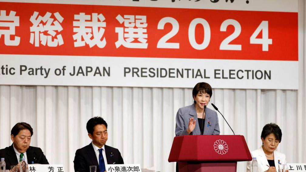 A woman in a suit speaks at a lectern in front of a sign saying '2024 presidential election' with Japanese characters. Three other people in suits, two men and a woman, sit alongside her.