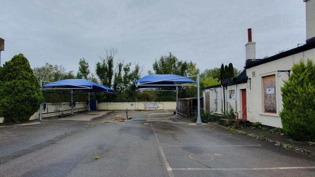 Blue canopies in an empty car park with low cream coloured walls and boarded up windows