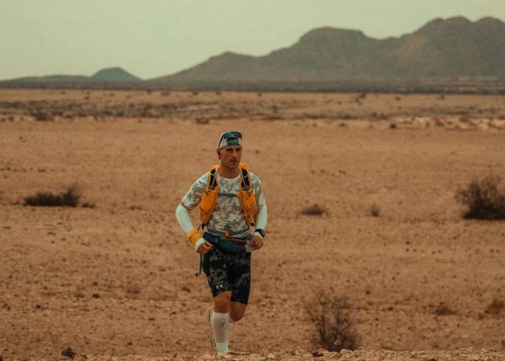 Jon Shield is wearing a hat running through the desert. He is near the bottom of the photo with a mountain landscape in the background.