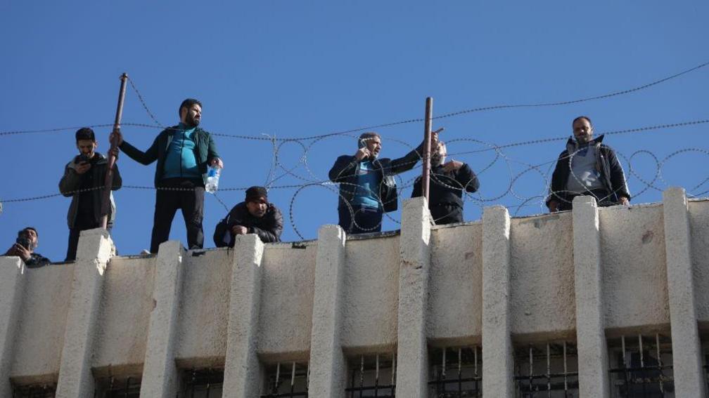 A group of men stand atop a concrete building with swirls of barbed wire around the edge, at Saydnaya prison, Syria