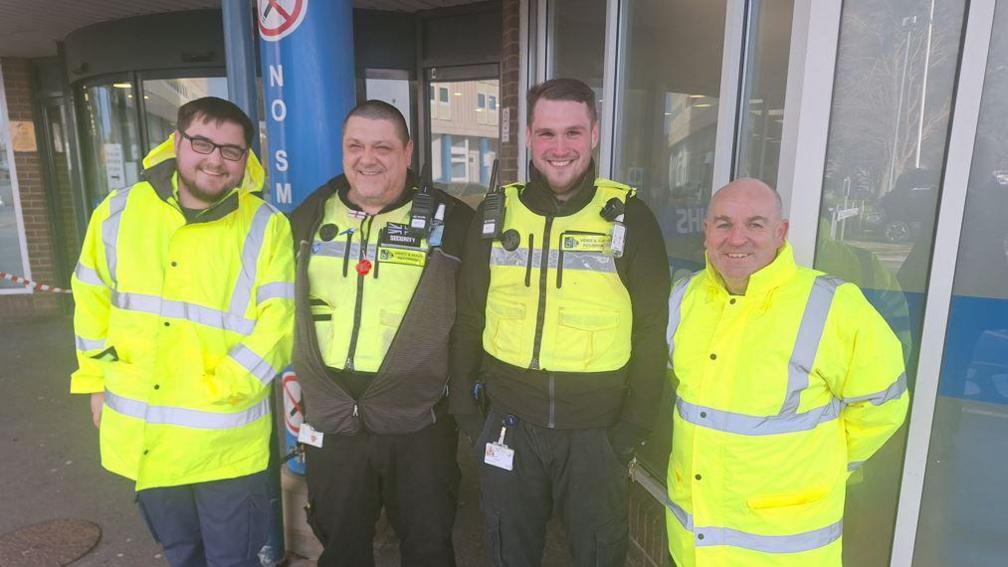 Maintenance assistant Luke Ferguson, security team members Gary Pickard and Aaron Sparrow, and plumber Ste Loughran smiling outside the University Hospital of North Tees. They are wearing high visibility clothing.