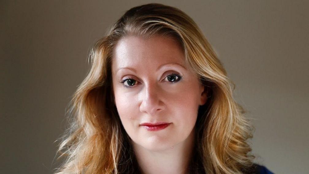 A woman with long dark blond hair looks into the camera with a neutral expression. 