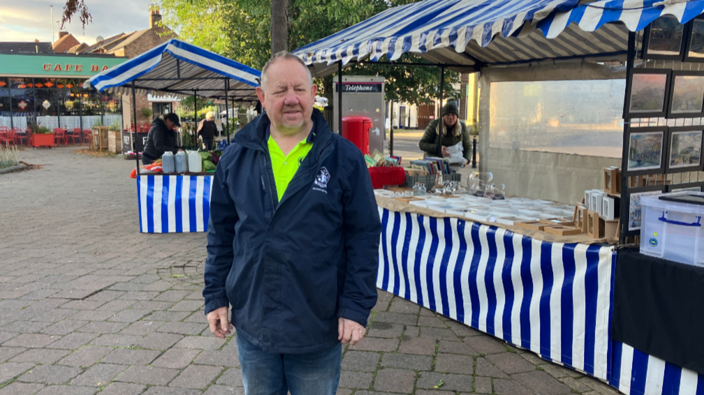 A man stood in front of a market stall in a blue jacket and with his hands by his sides as he looks to the camera