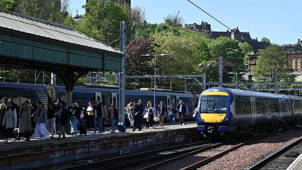A ScotRail train arrives at Waverly station in Edinburgh in the sunshine, the castle in the background. People are making their way off the train and up the platform.
