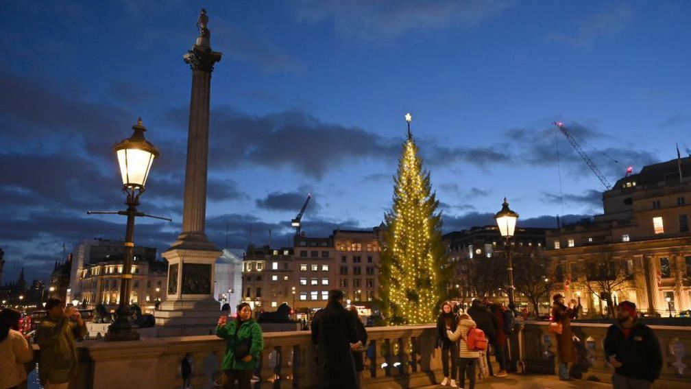 Trafalgar Square in 2023 - showing Christmas tree and people taking pictures nearby at night time 