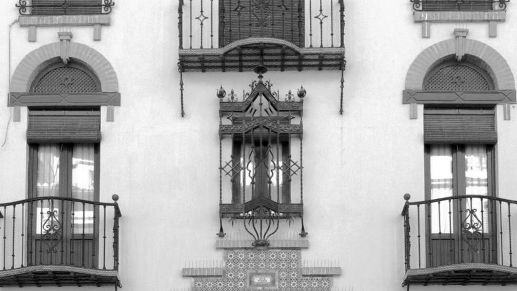 A black and white image of a building with intricate architecture. There are two long doors either side of a smaller window with bars over the top. Each door has a small veranda. 