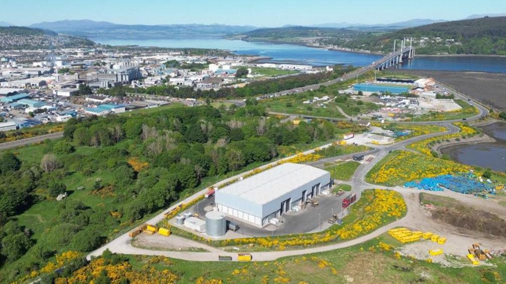 An aerial view of Inverness, looking back on the city from the East Longman area. A large grey rectangular building is surrounded by grass and trees near the bottom of the image. The Kessock bridge can be seen in the far right-hand side of the picture with the city centre in the far left. It's a sunny day and the Beauly Firth is bright blue.